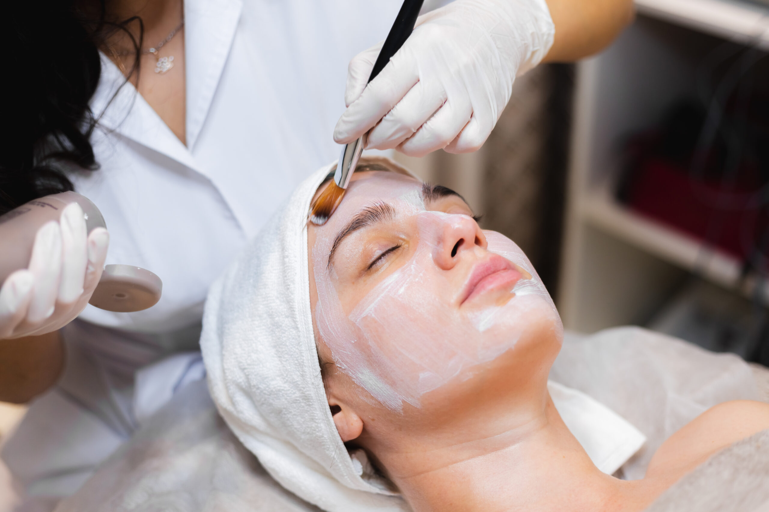 beautician with a brush applies a white moisturizing mask to the face of a young girl client in a spa beauty salon